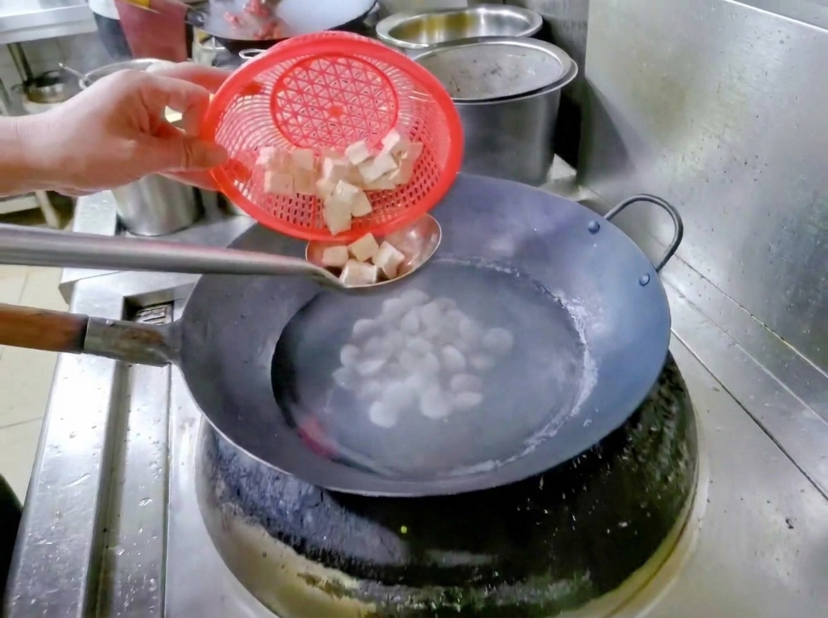 Una mano che regge un cestino scolapasta rosso, facendo cadere tofu a cubetti in un wok di acqua e vongole.