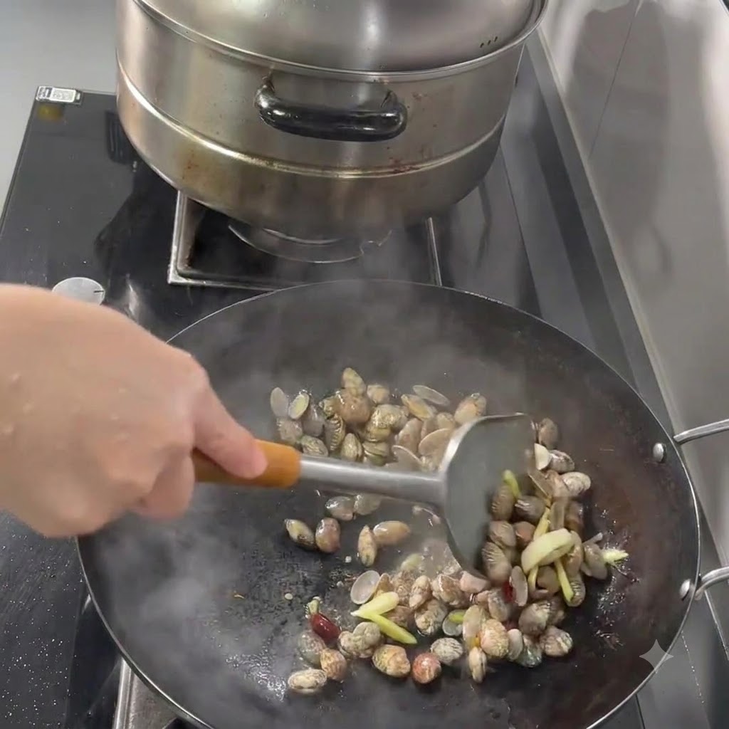 Vongole, aglio, zenzero e peperoncini saltati in un wok con una spatola di metallo.