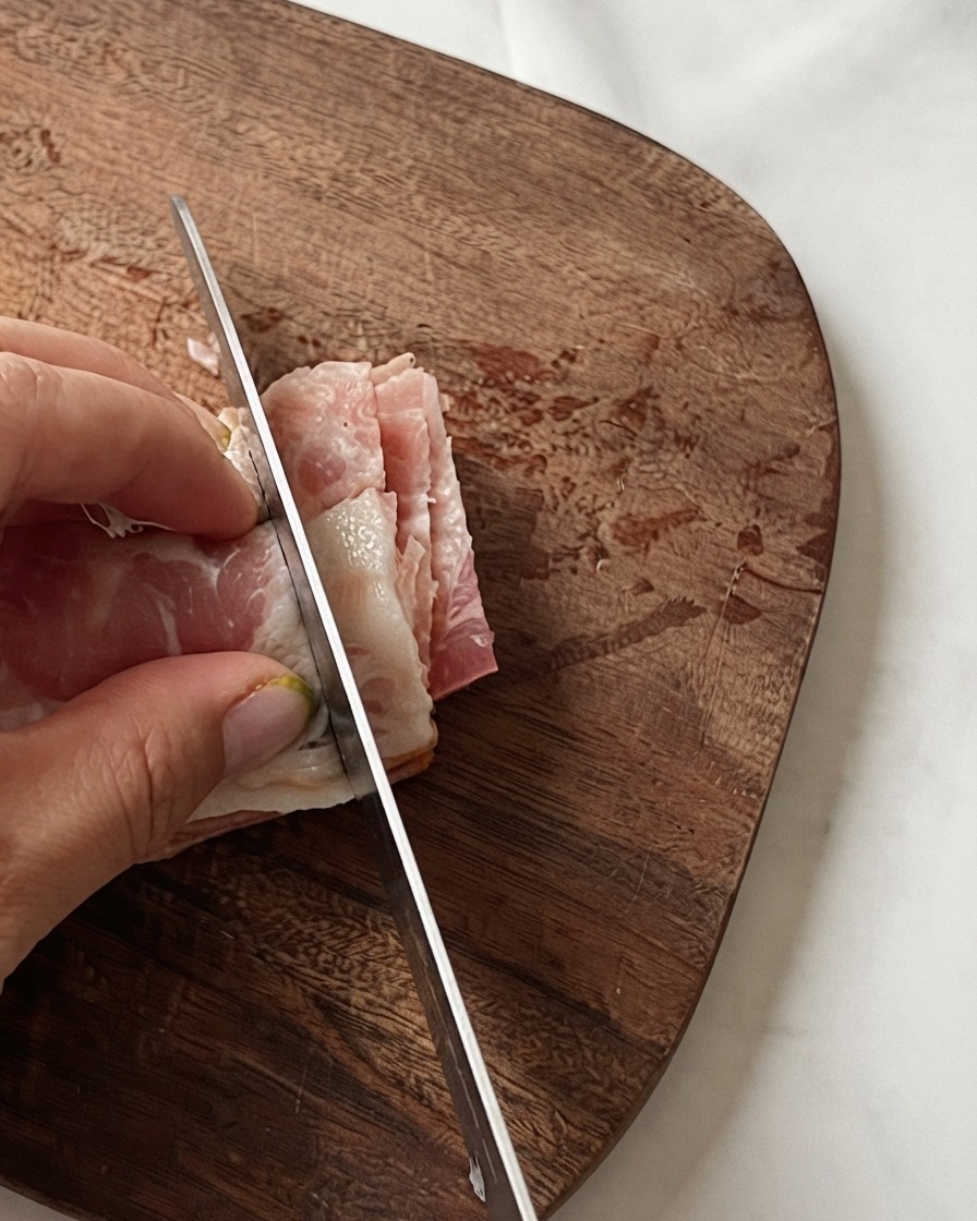 La mano di uno chef che tiene strisce di pancetta impilate mentre un coltello le taglia a piccoli pezzi su un tagliere di legno.
