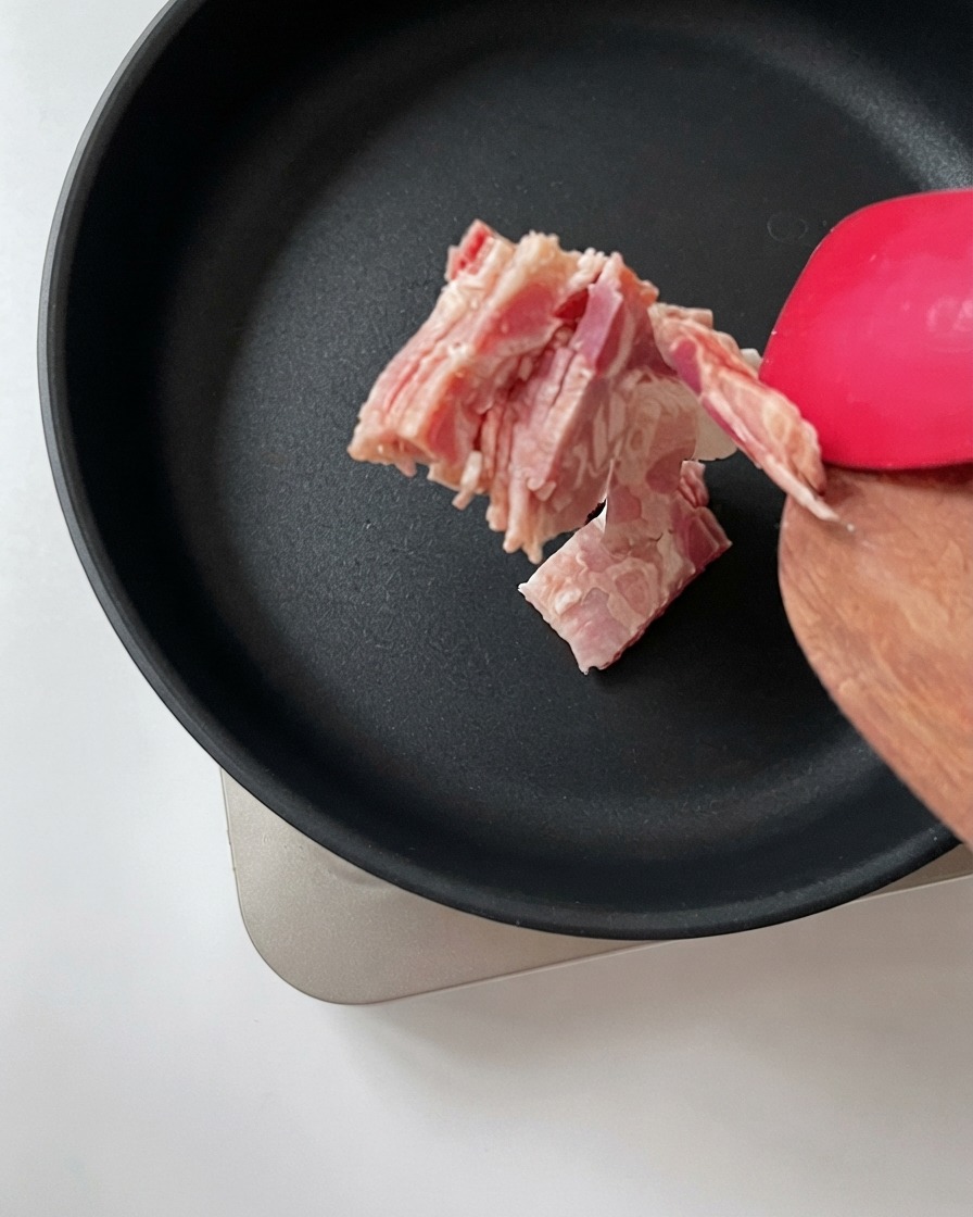 Pancetta sminuzzata trasferita da una spatola di legno in una padella nera calda.