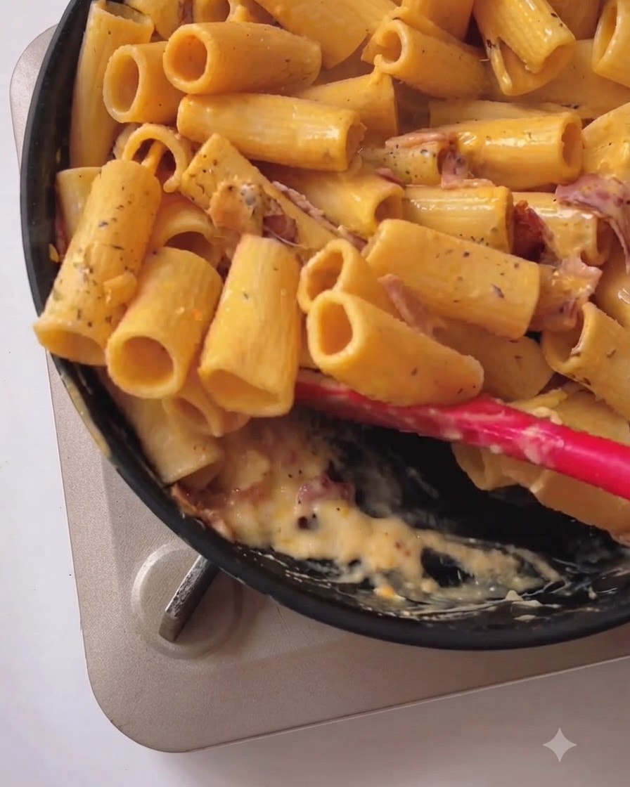 Pasta rigatoni mescolata e amalgamata con salsa cremosa gialla e pancetta in una padella nera usando una spatola in silicone rosso.