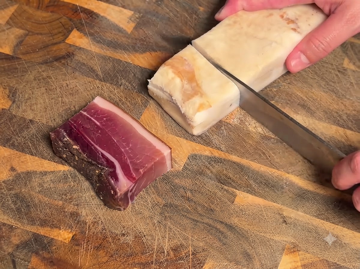 Un coltello che affetta un blocco di carne stagionata e pancetta su un tagliere di legno.