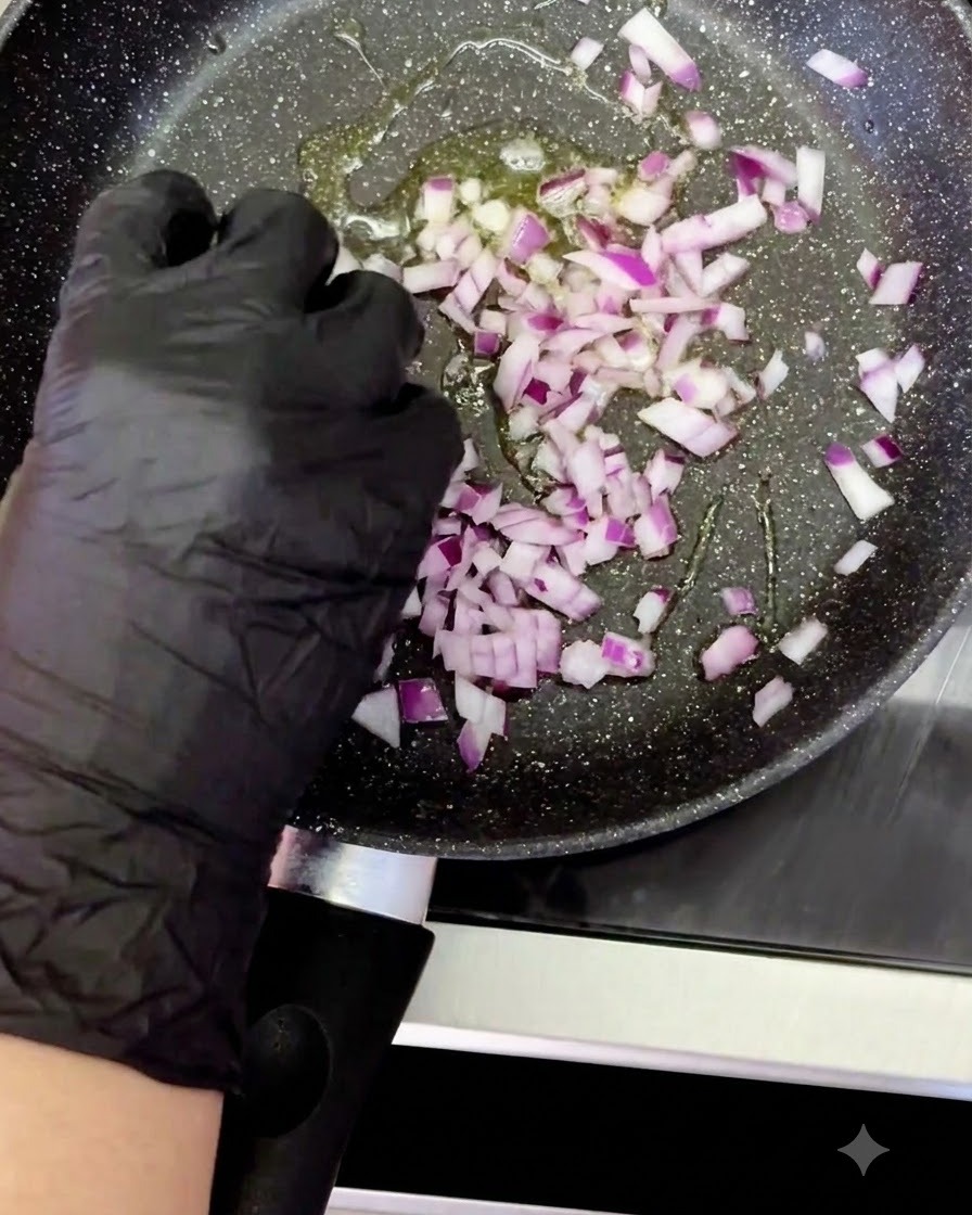 Una mano guantata che aggiunge cipolla rossa a dadini in una padella riscaldata con olio sfrigolante.