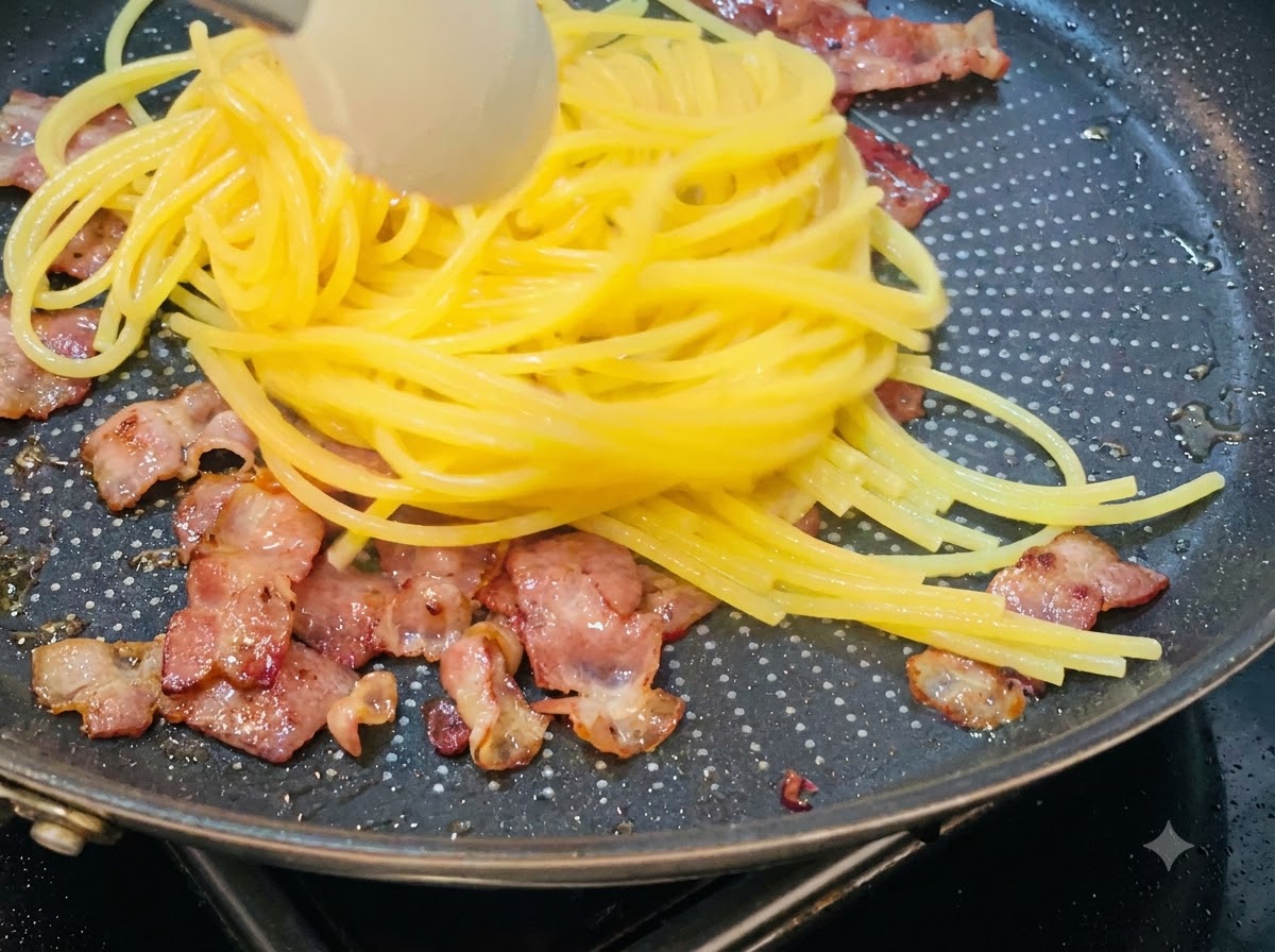 Spaghetti cotti sollevati e trasferiti in una padella contenente pezzi di pancetta fritta.