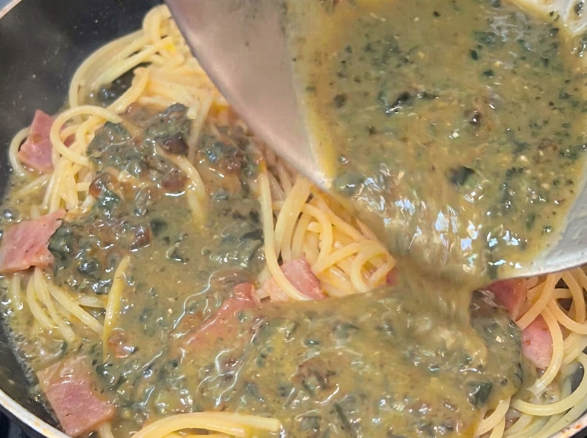 Salsa cremosa e punteggiata di uova centenarie versata da una ciotola sopra spaghetti caldi e pancetta in una padella.