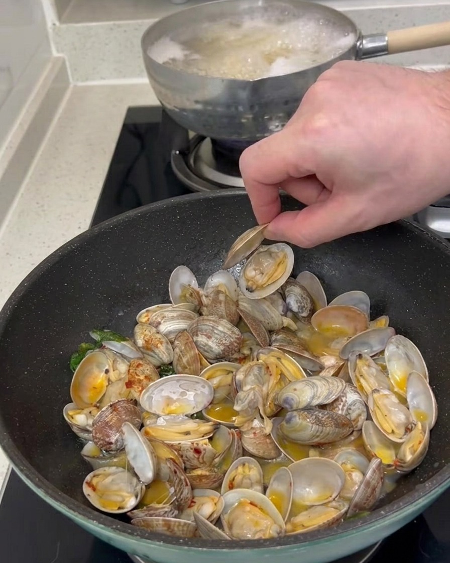 La mano di una persona che raccoglie un guscio di vongola aperto da una padella piena di vongole cotte e sugo.