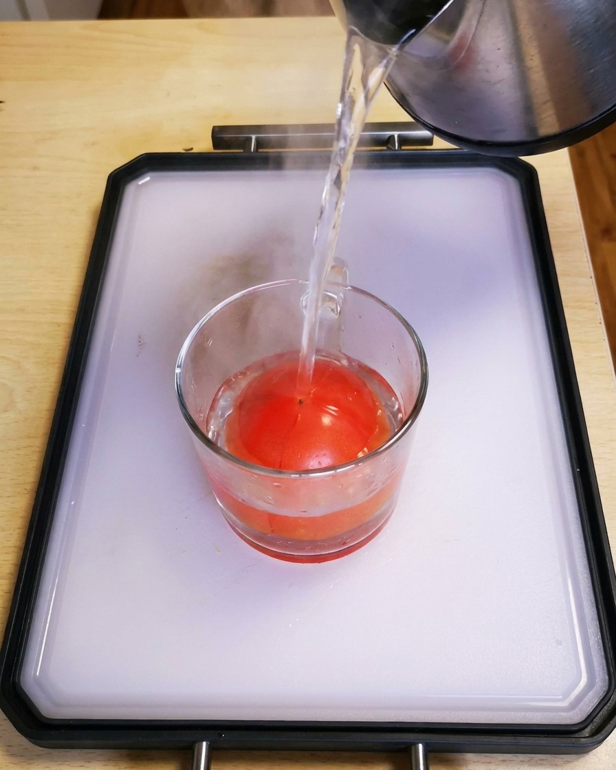 Acqua calda versata da un bollitore in un bicchiere di vetro contenente un pomodoro rosso fresco.