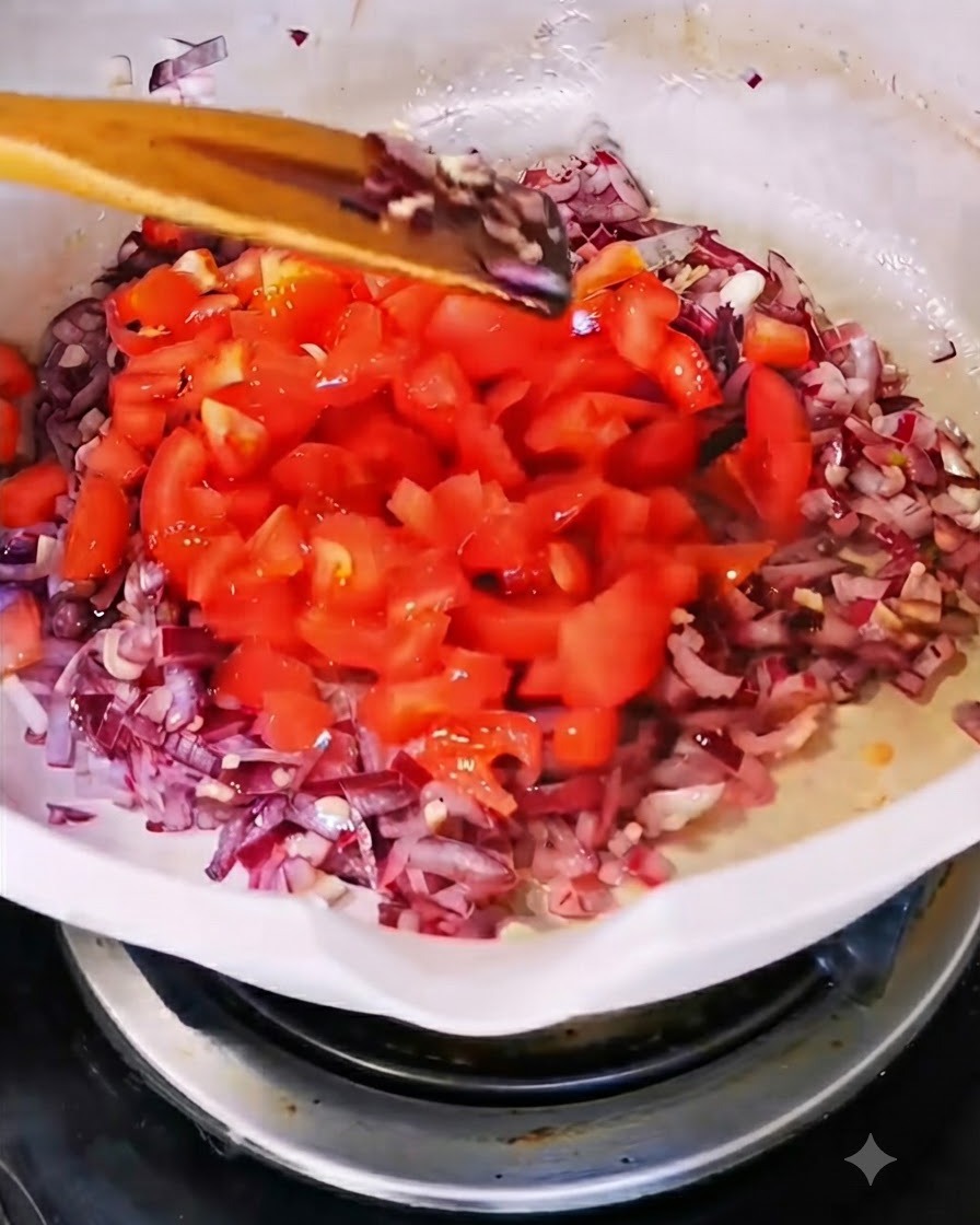 Pomodori rossi tagliati a cubetti aggiunti in una padella bianca contenente cipolla e aglio rosolati.