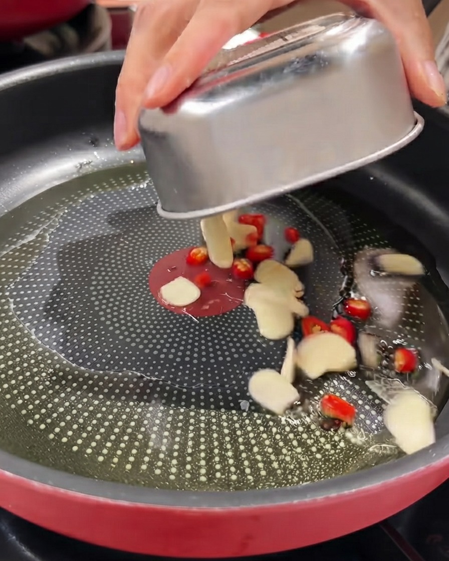 Aglio affettato, peperoncini rossi ed erba cipollina versati da un contenitore di metallo in una padella oliata.