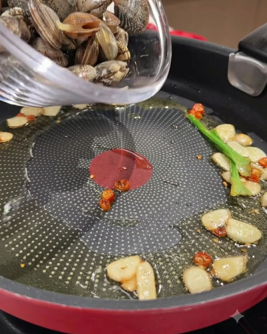 Vongole fresche versate da una ciotola di vetro in una padella con aglio, peperoncini e olio.