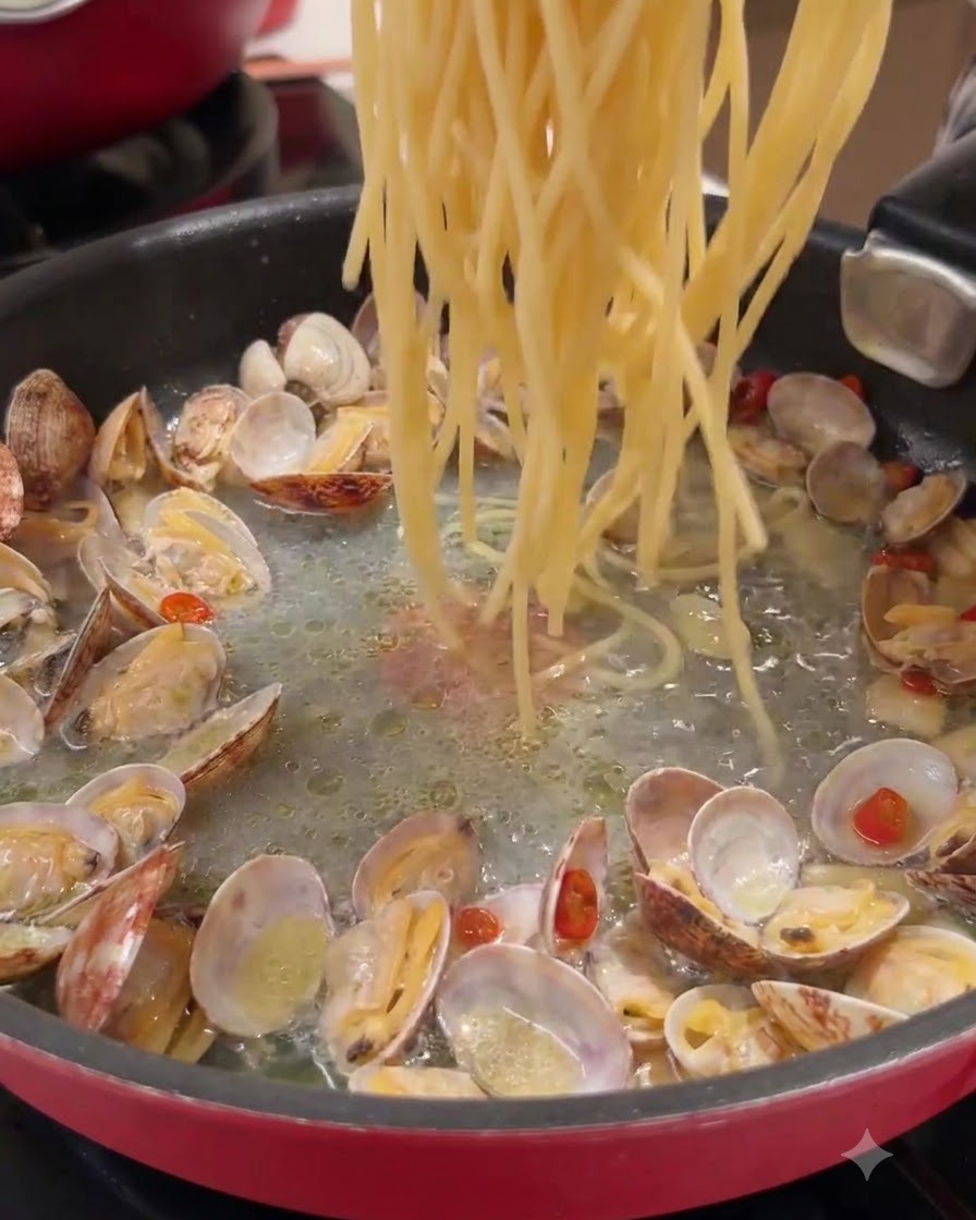 Lunghi fili di pasta gettati in una padella contenente vongole cotte e liquido condito.