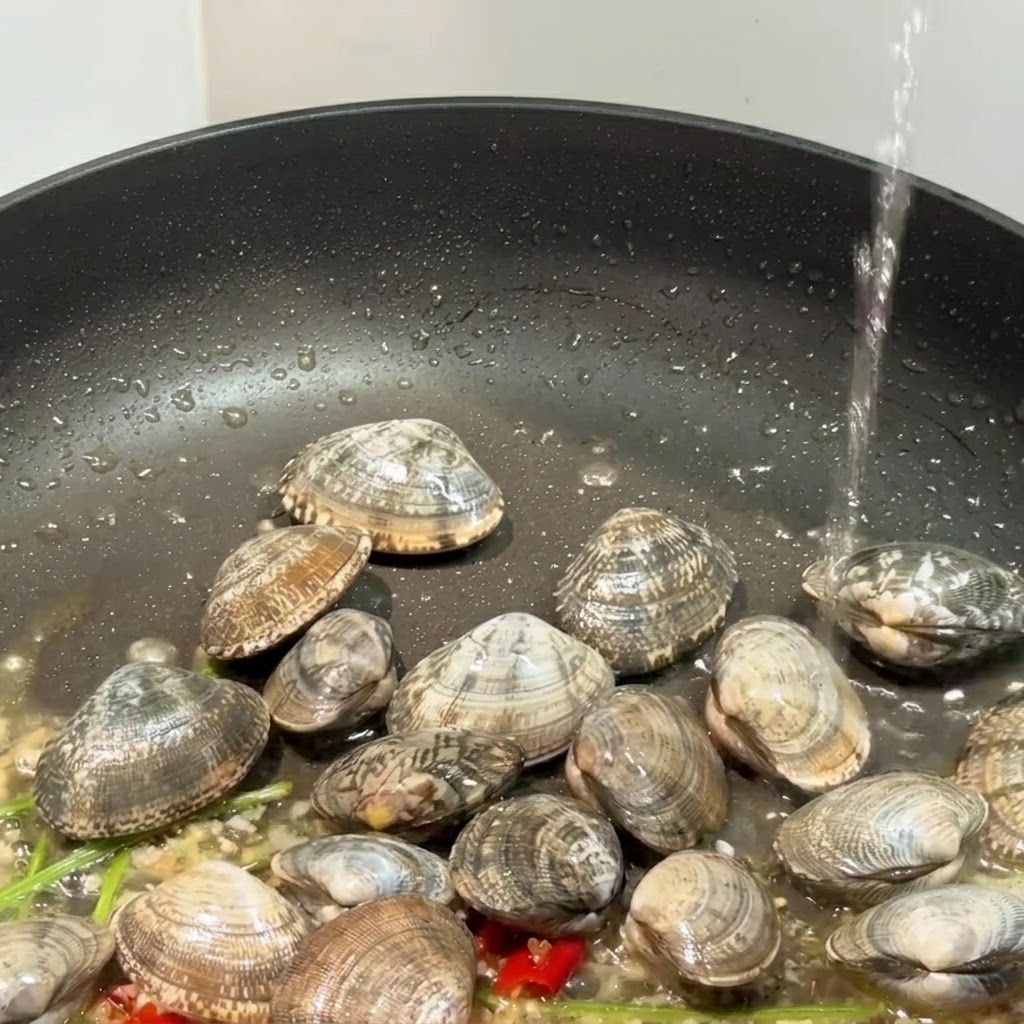 Una padella nera contenente vongole, aglio e peperoncini con liquido versato allinterno.