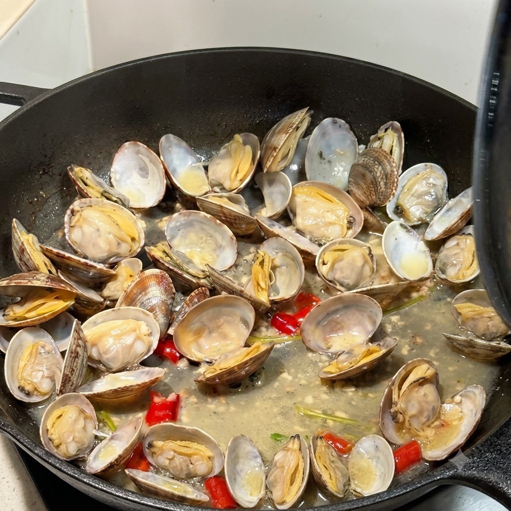 Vongole aperte che cuociono nel brodo dentro una padella con coperchio parzialmente visibile.