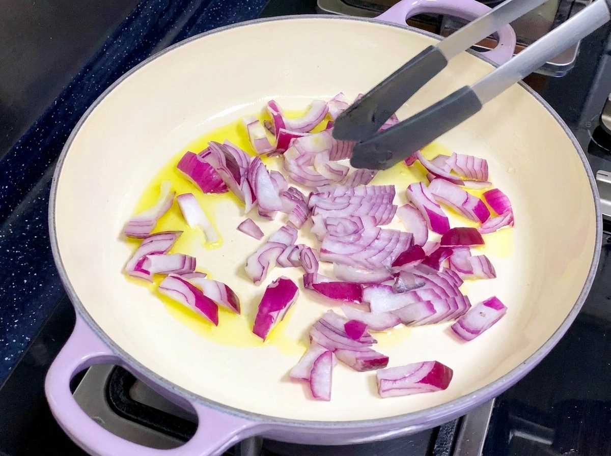 Una padella di colore chiaro contenente olio doliva e cipolle rosse tritate mescolate con le pinze.