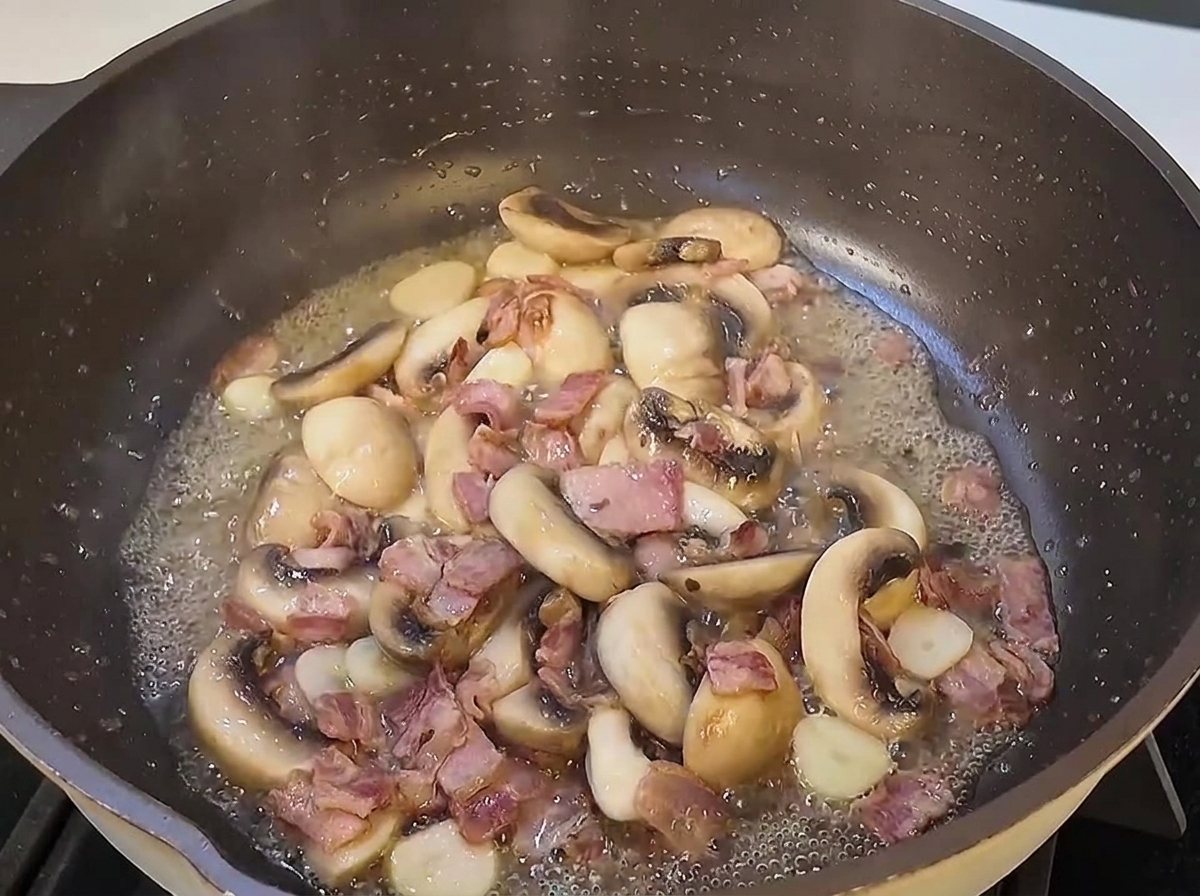 Spaghetti cotti che vengono saltati in una padella capiente con fette di funghi e pezzi di pancetta.