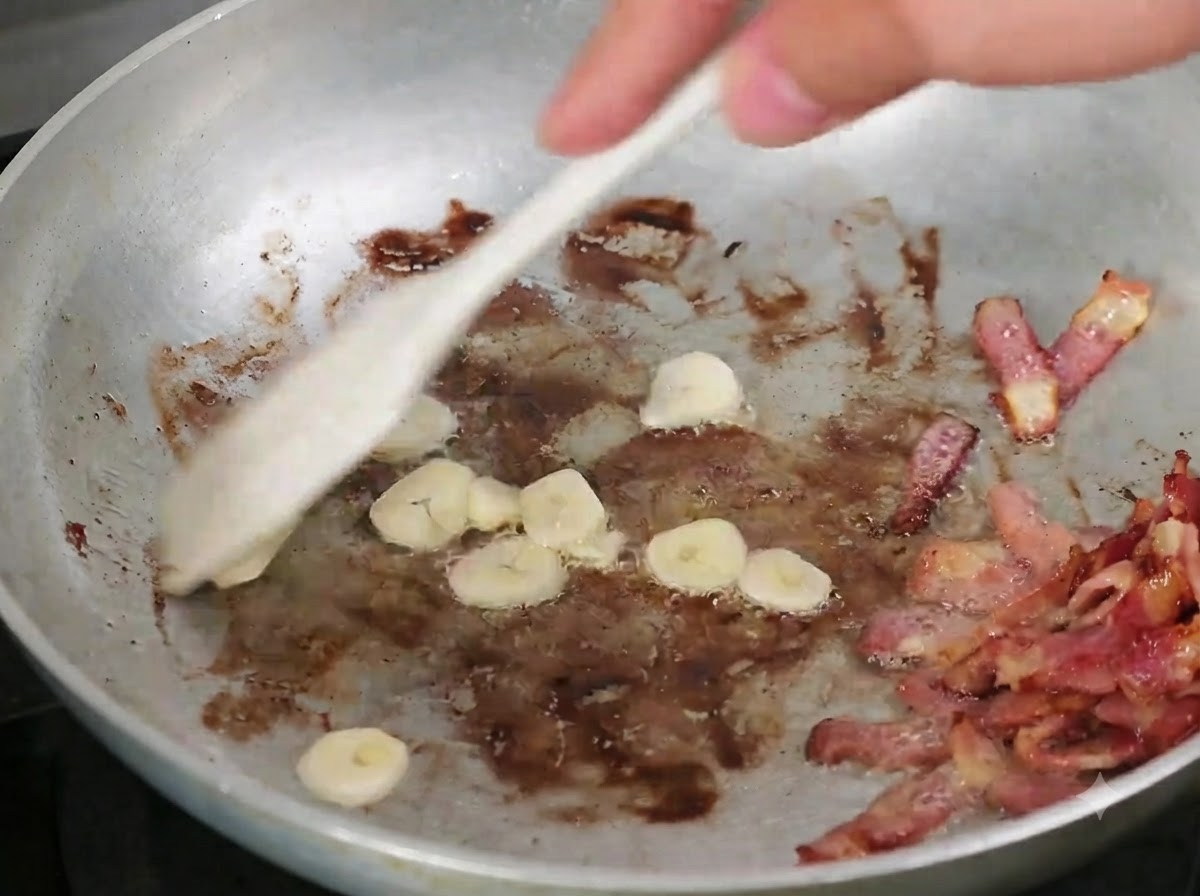Aglio a fette mescolato nella padella con la pancetta rosolata utilizzando un cucchiaio di legno.