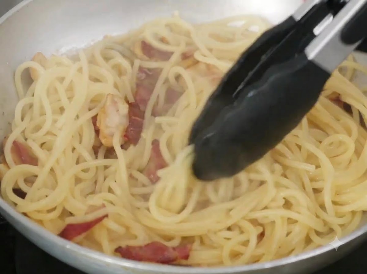 Burro e acqua di cottura mescolati agli spaghetti in una padella usando delle pinze.