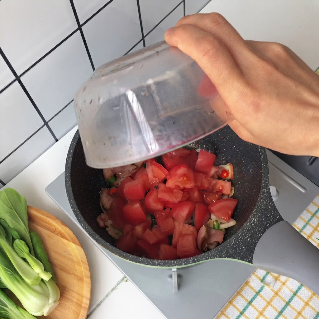 Mano che versa i pomodori tritati da una ciotola di vetro in una padella contenente pancetta fritta e cipollotti.