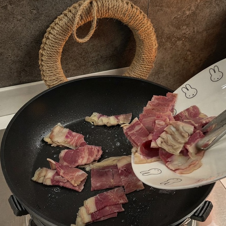 Pezzi di pancetta affettata aggiunti e saltati in una padella nera.