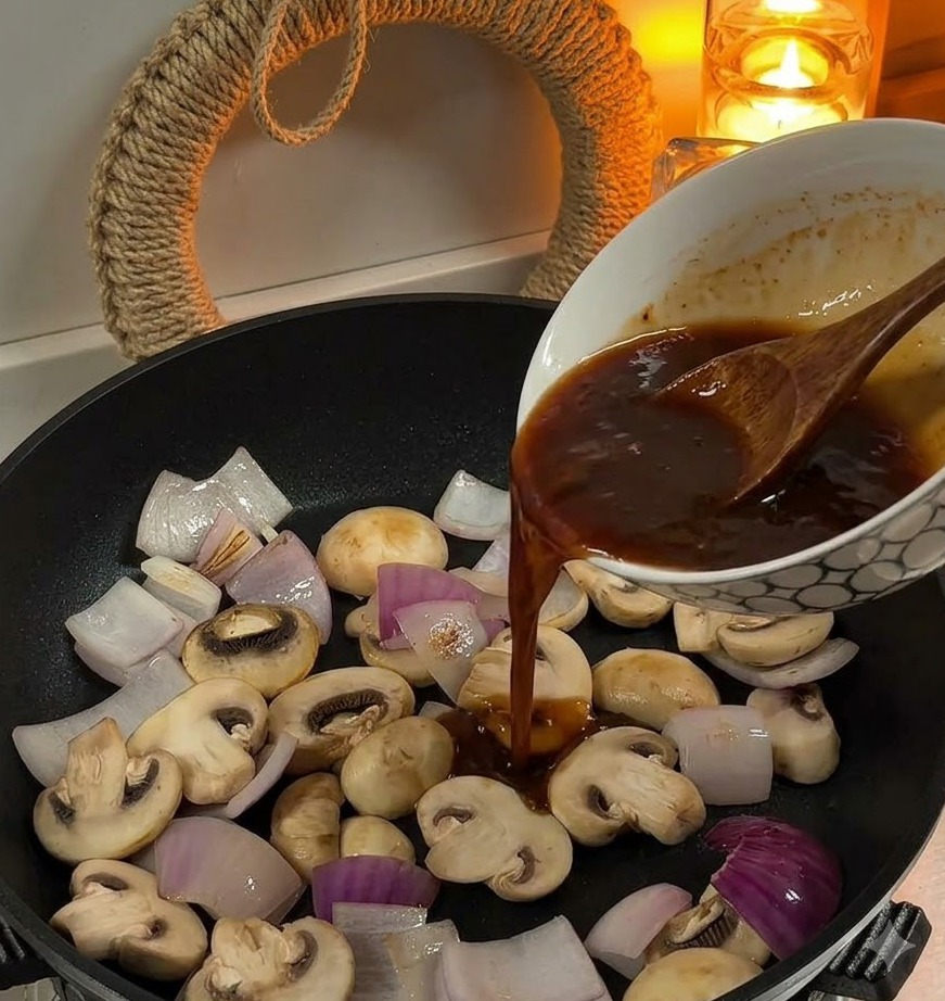 Versamento di salsa marrone da una ciotola in una padella contenente cipolle affettate e funghi bianchi.