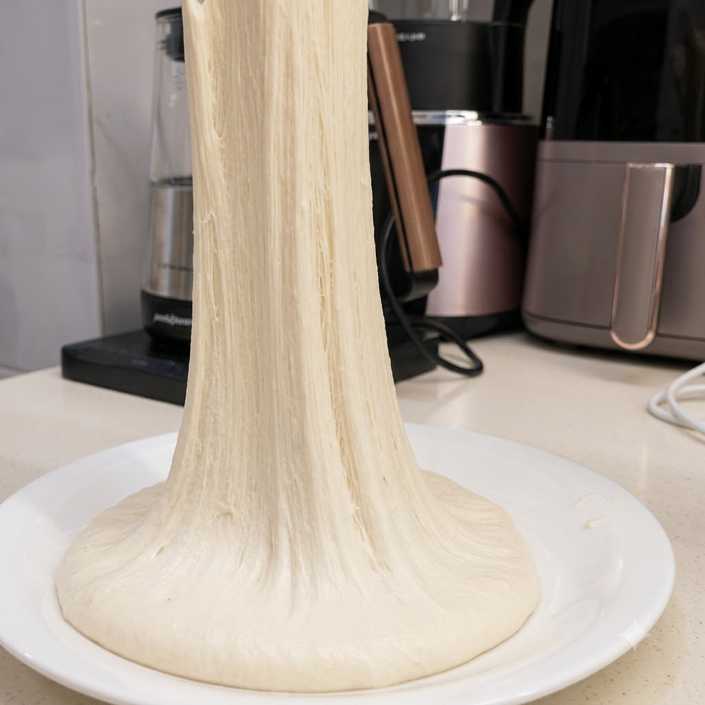 Un filo lungo e continuo di impasto per pizza altamente elastico tirato verso lalto da un piatto bianco.