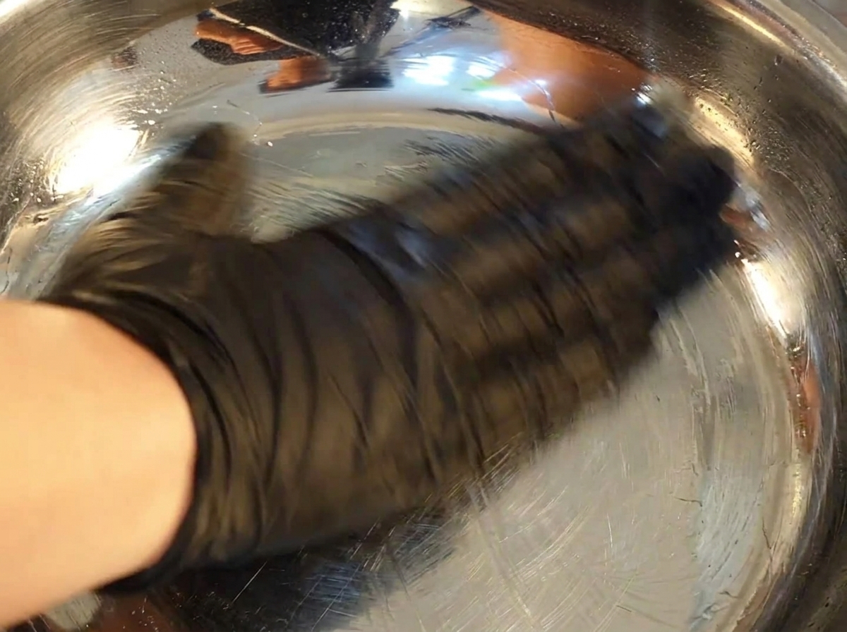 Una mano con un guanto nero usa un tovagliolo di carta per ungere linterno di una ciotola di acciaio inossidabile con olio.