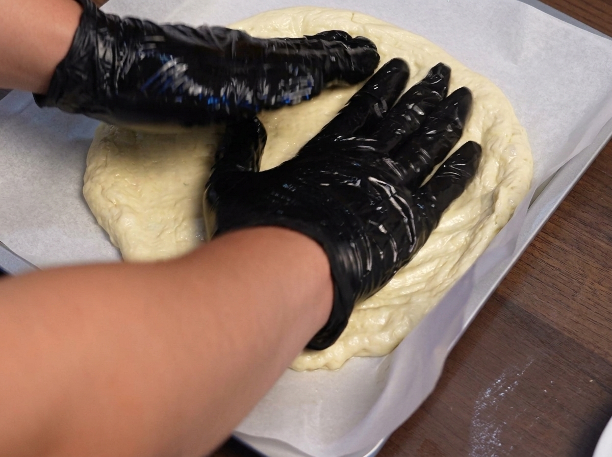 Mani guantate che modellano limpasto della pizza verso lesterno su una teglia foderata di carta forno.