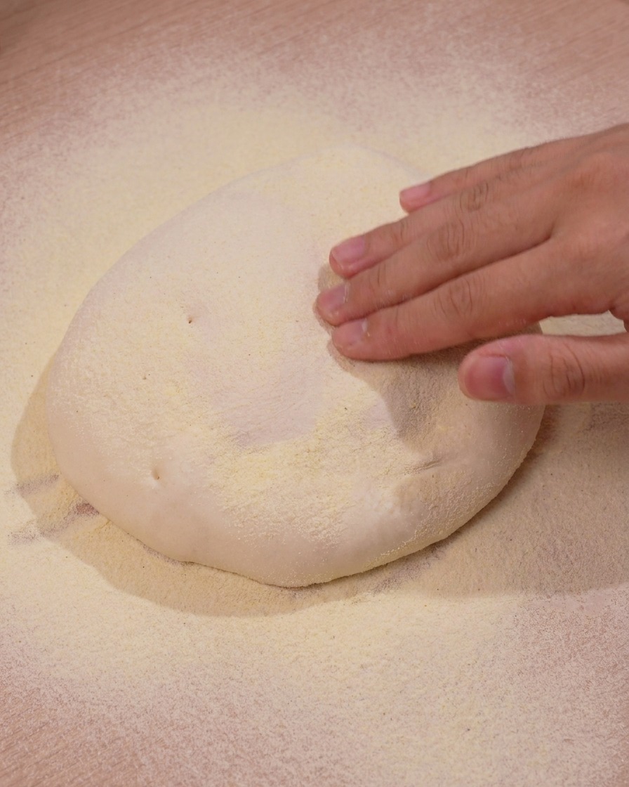 Mano che preme delicatamente verso il basso su un mucchio di impasto per pizza poggiato su farina di granoturco gialla.