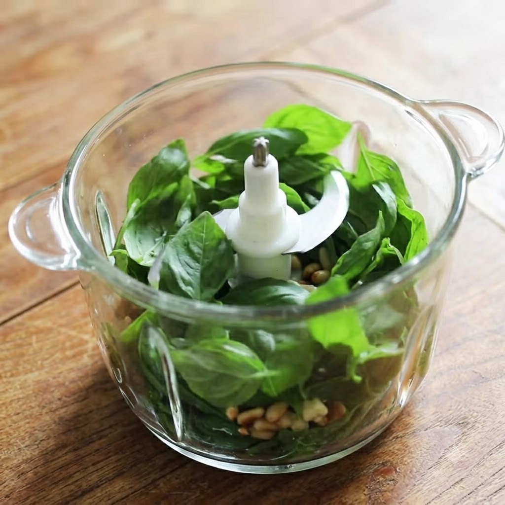 Un contenitore di un frullatore in vetro riempito con foglie di basilico fresco, pinoli, aglio e formaggio pronti per essere frullati.