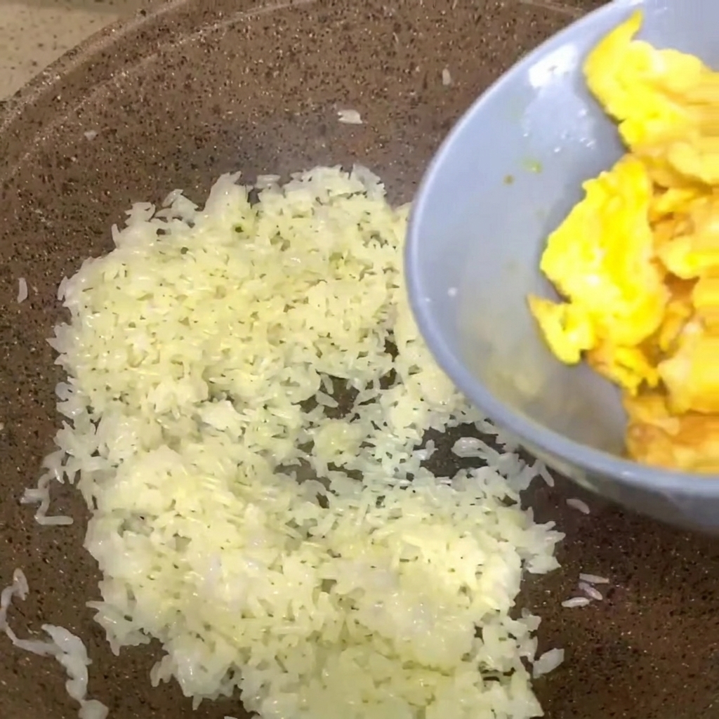 Uova strapazzate da una ciotola grigia versate in una padella di riso bianco cotto.