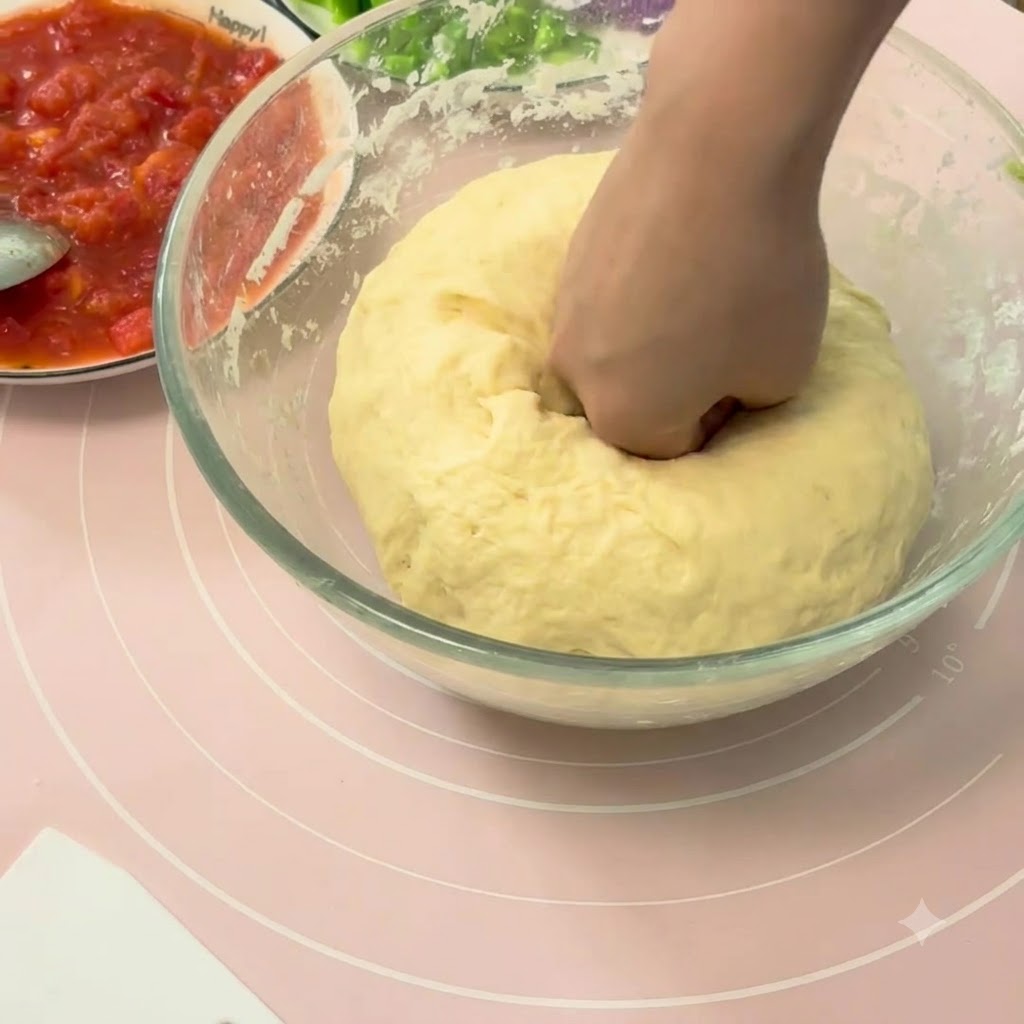 Una mano che schiaccia verso il basso una grande palla di impasto per pizza lievitato dentro una ciotola di vetro.