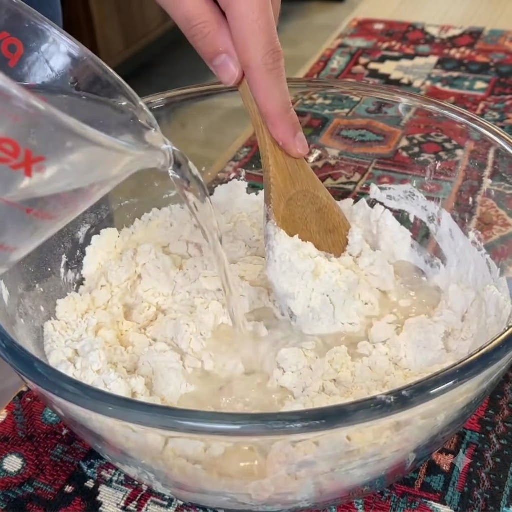 Acqua versata da un misuratore in una ciotola di vetro di farina mentre un cucchiaio di legno mescola.