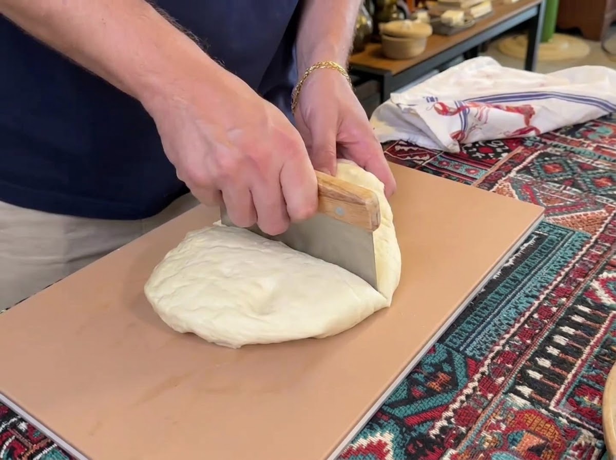 Un tarocco per pasta che taglia un grande mucchio di impasto bianco per pizza in quattro pezzi separati su un tagliere.