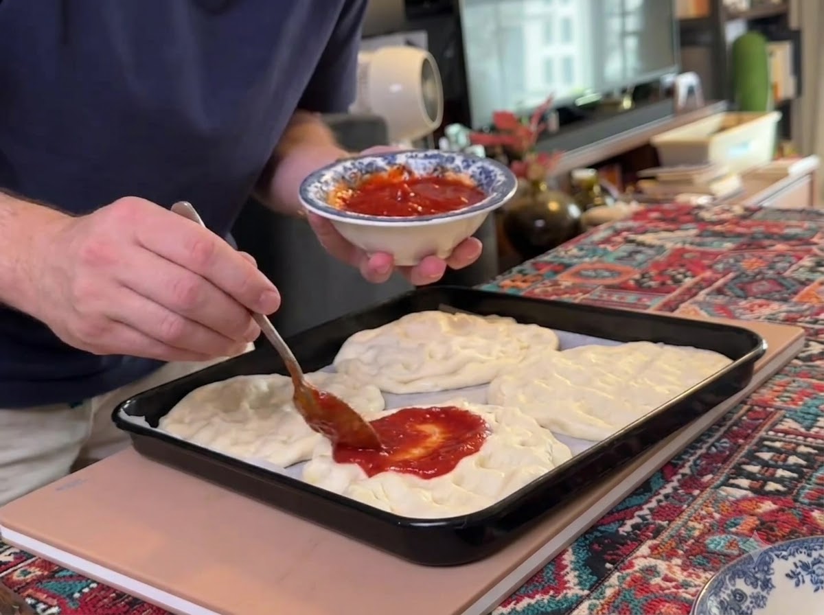 Un cucchiaio che distribuisce la salsa di pomodoro rossa su più basi di pasta per pizza appiattite disposte su una teglia nera.