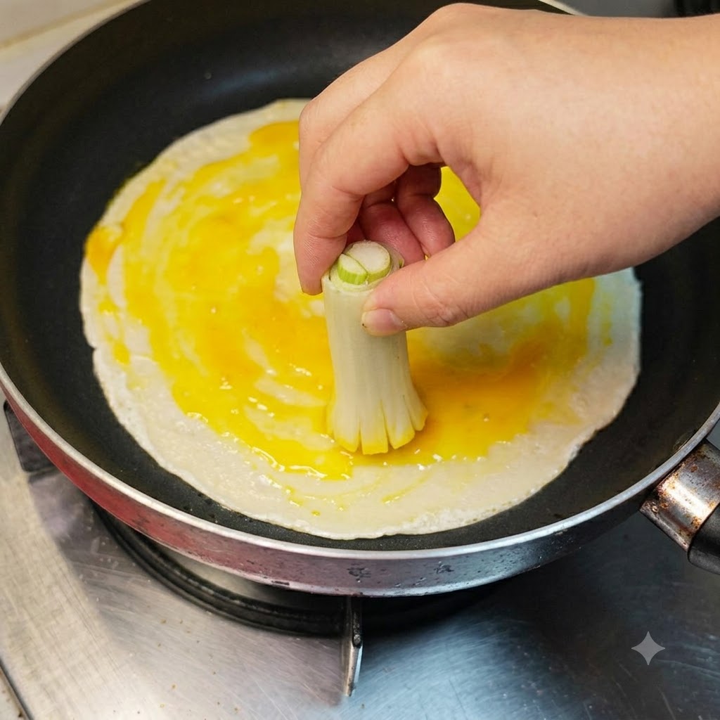 Una mano che usa un mazzetto di gambi di cipollotto legati per spennellare lalbume e il tuorlo duovo rotto su un pancake.