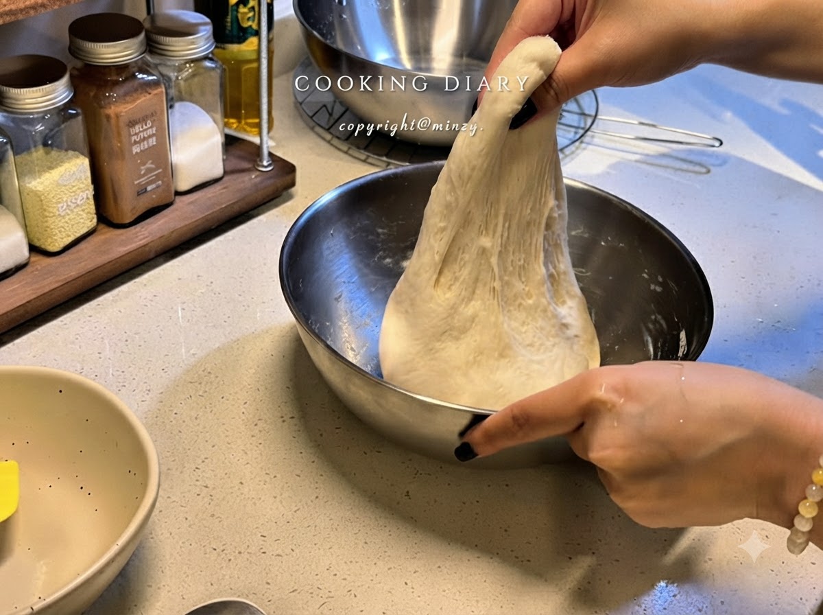 Un paio di mani sollevano una porzione di impasto elastico per pizza verso lalto allinterno di una ciotola di metallo per allungarlo.