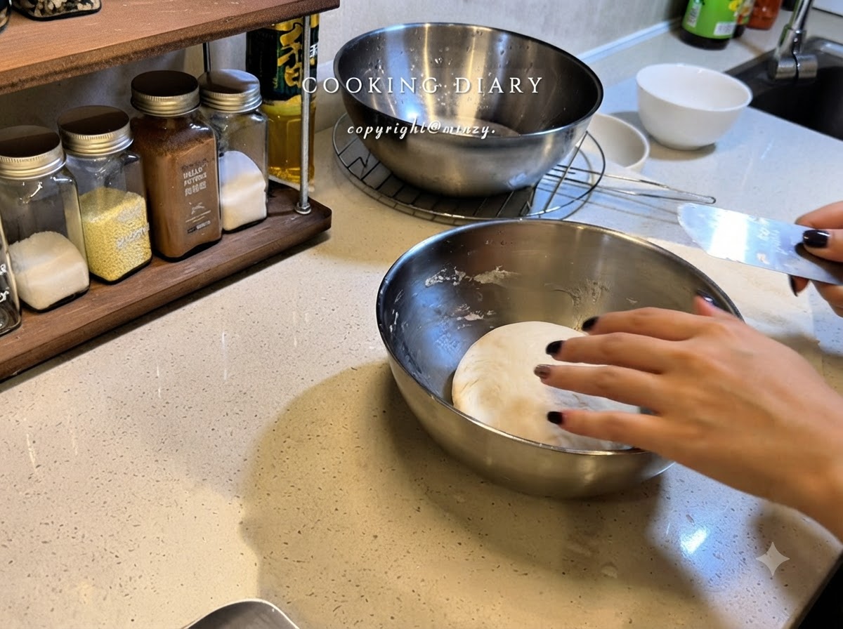 Una mano che stende la pellicola trasparente su una ciotola in acciaio inossidabile contenente una pallina rotonda di impasto per pizza.