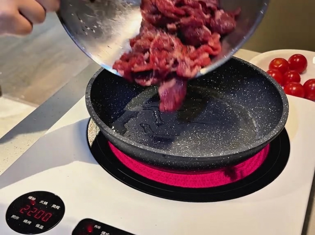 Fettine di manzo crudo marinato versate da una ciotola di metallo in una padella maculata calda su un piano cottura a induzione.