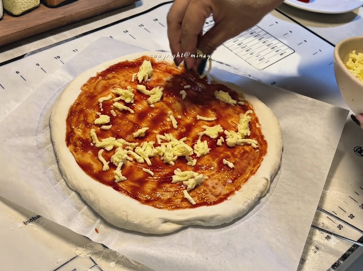 Mani che cospargono formaggio grattugiato su una base per pizza ricoperta di salsa di pomodoro rossa.
