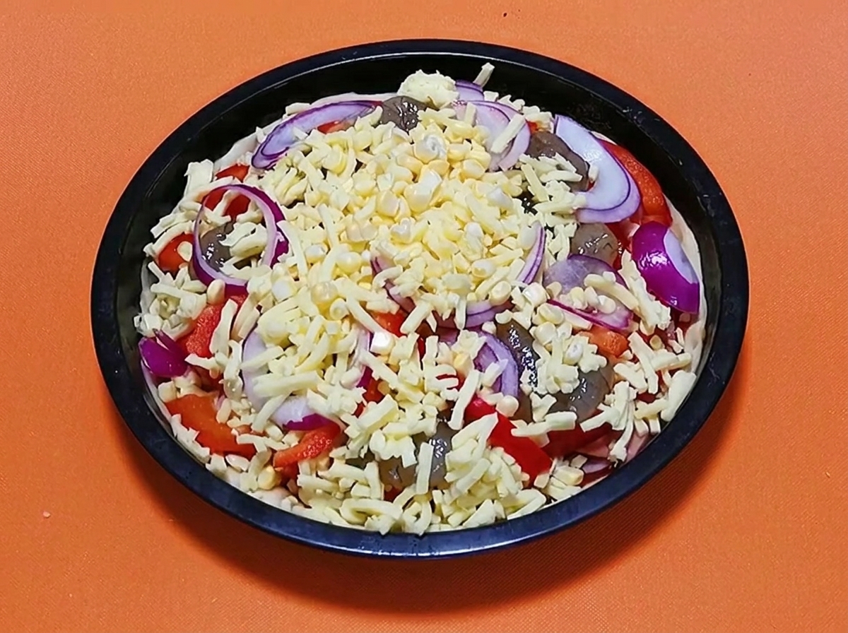 Pizza di mare cruda completamente assemblata guarnita con un ultimo strato di formaggio grattugiato e fette di cipolla rossa, pronta per essere infornata.
