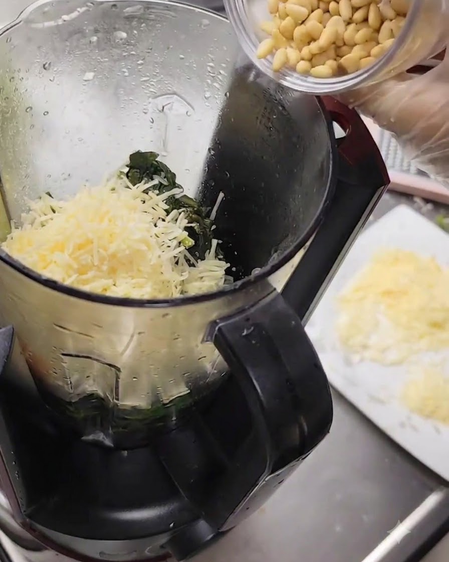 Pinoli versati da una piccola tazza nel frullatore pieno di formaggio e basilico.