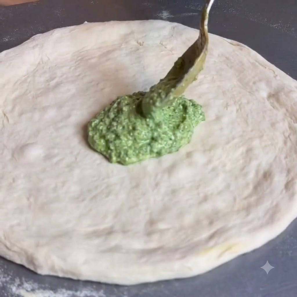 Un cucchiaio che distribuisce la salsa di pesto verde al centro di un disco di impasto per pizza.