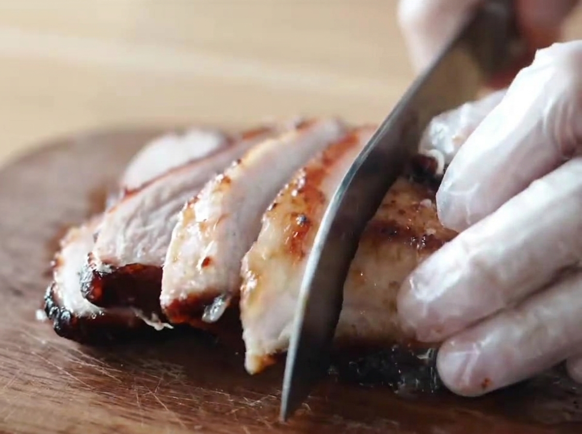 Un pezzo di chashu di maiale caramellato e glassato al miele tagliato a fette spesse da una persona con i guanti su un tagliere di legno.