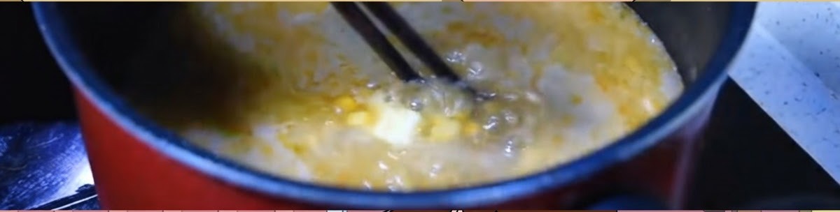Bacchette che mescolano un brodo di miso arancione in forte ebollizione con mais e una noce di burro in una pentola scura.