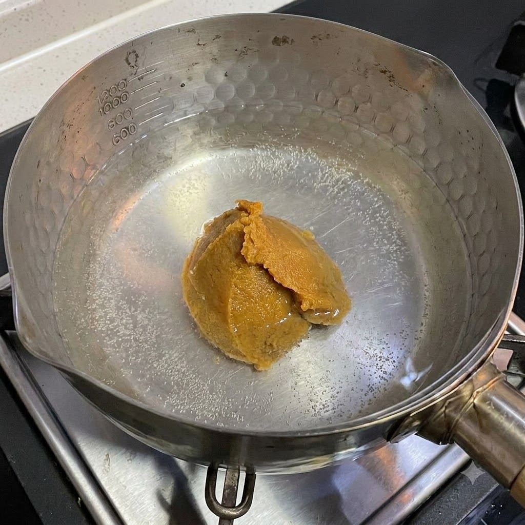 Un cucchiaio di pasta di miso bianco che si scioglie in una pentola di acqua calda sul piano cottura.