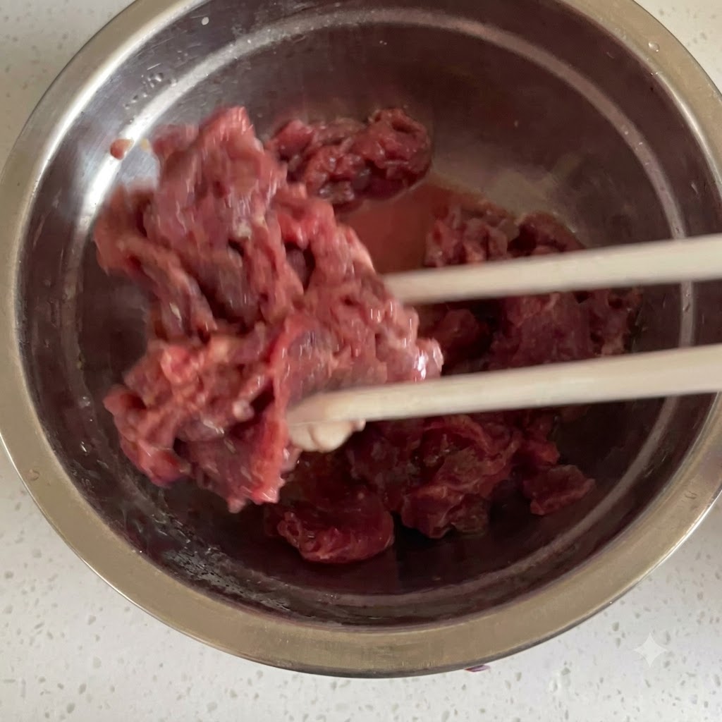 Manzo crudo tagliato a fettine sottili mescolato accuratamente con gli ingredienti della marinata usando le bacchette allinterno di una ciotola in acciaio inossidabile.