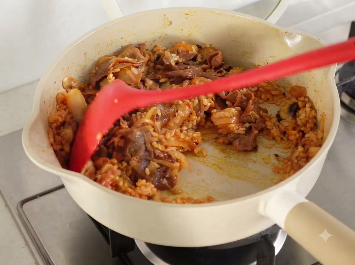 Tutti gli ingredienti e il riso saltati accuratamente insieme fino a essere rivestiti di succo di kimchi in una padella.