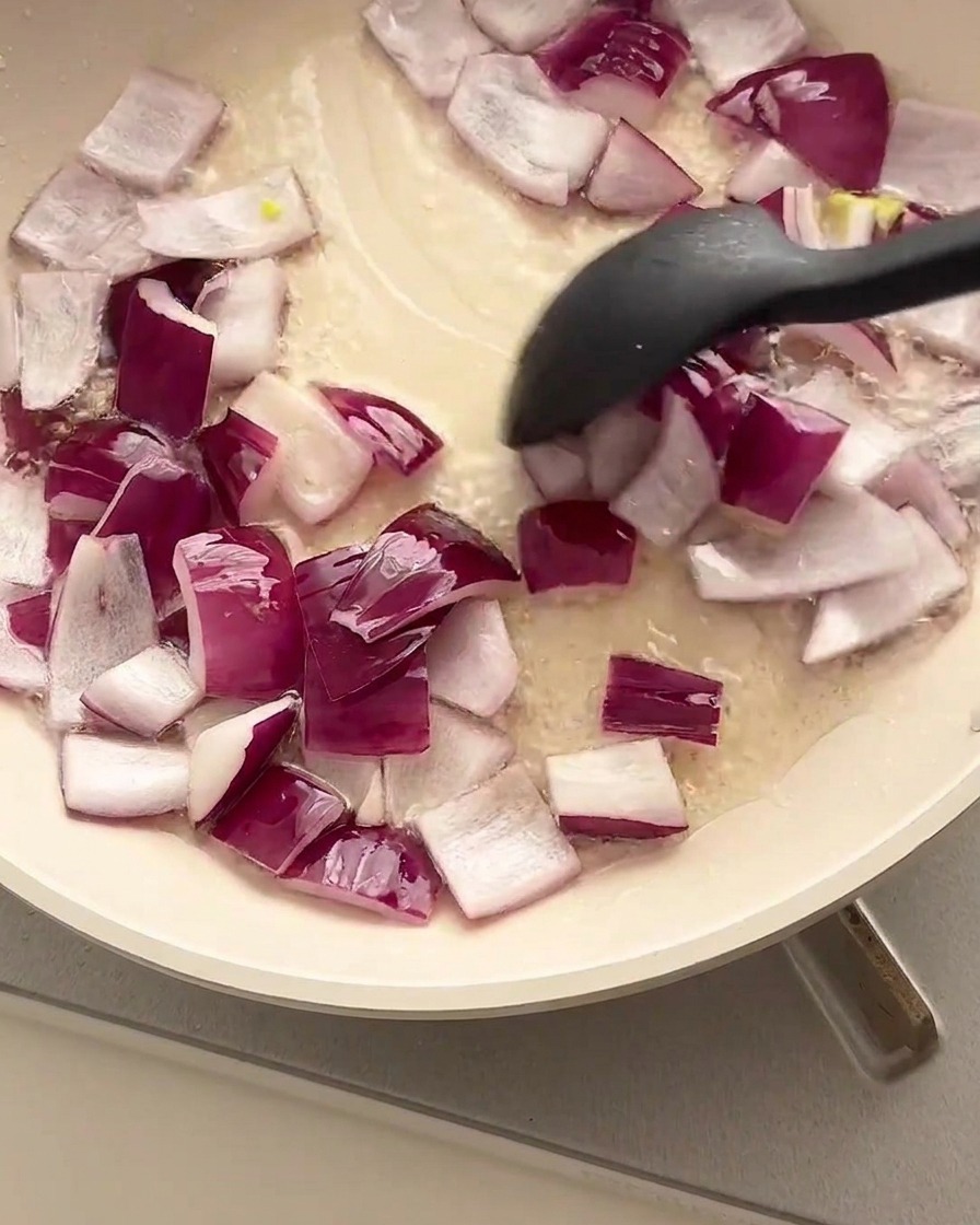 Cipolle rosse a cubetti saltate in una padella oliata usando una spatola nera.