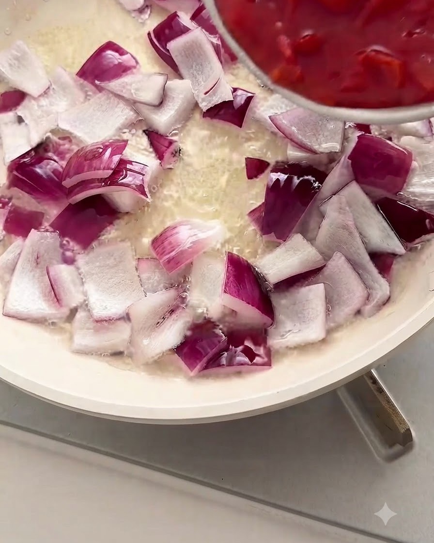 Pomodori rossi a pezzetti versati da una ciotola in una padella bianca piena di cipolle rosse a cubetti e olio.