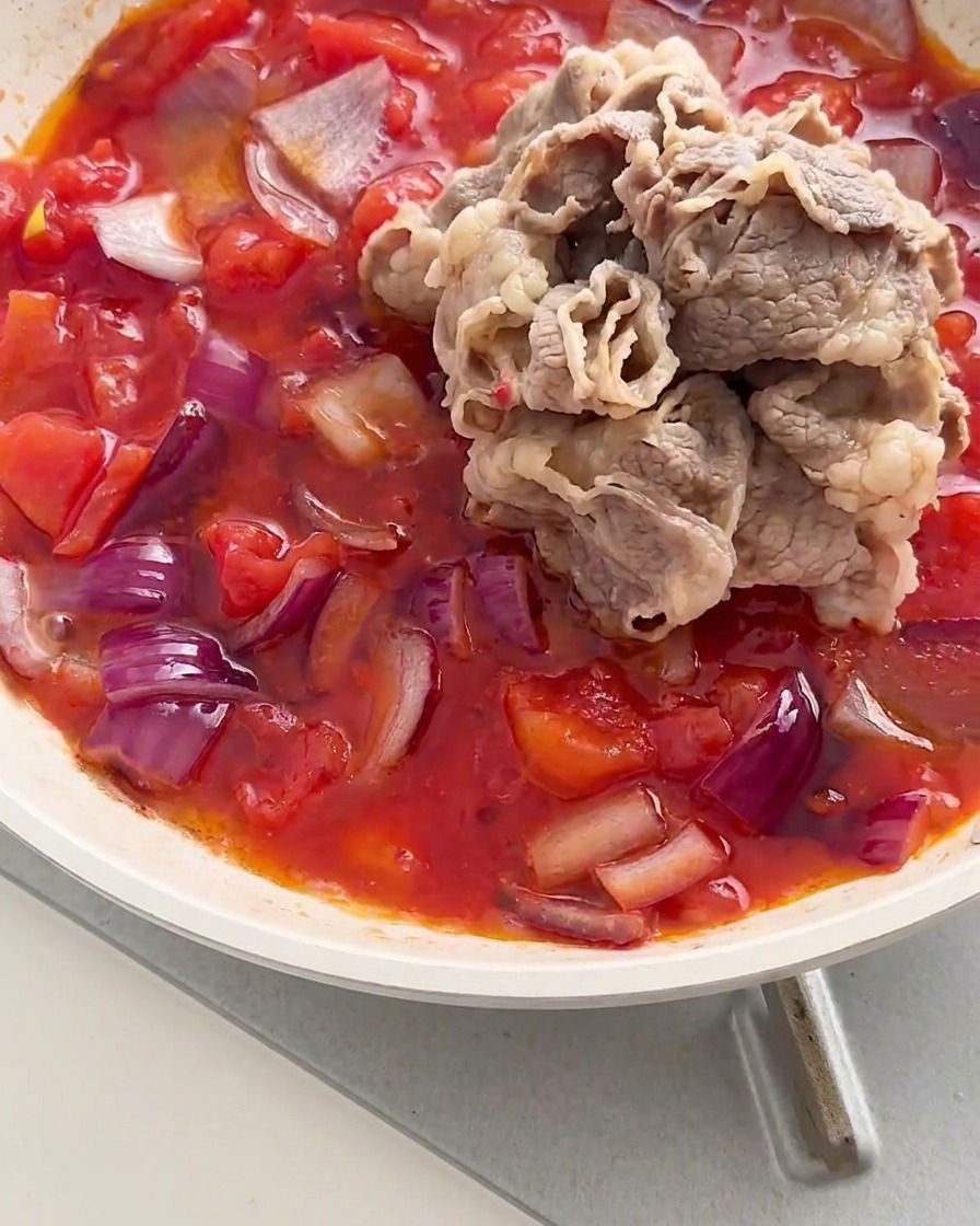 Un mucchio di involtini di manzo sbollentati aggiunti in una padella con un composto sobbollente di pomodori rossi e cipolle.